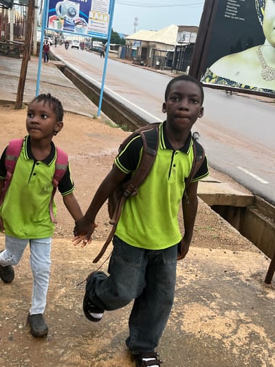 In Sabojibi, Nigeria, two children, a boy and a younger girl, stand together. The boy, positioned in the foreground and slightly higher than the girl, wears a bright lime green polo shirt with black accents on the collar and sleeves, and dark blue jeans. He carries a brown backpack. The girl, standing to his right and slightly lower, also sports a lime green polo shirt, this one with pink accents on the shoulders, and light blue jeans. She too has a backpack. They are holding hands, suggesting a connection or shared activity.

They are standing on a dirt and concrete surface, likely a sidewalk or roadside area, with a paved road visible behind them. The road appears wet, hinting at recent rain or ongoing damp conditions. The sky above is overcast, suggesting a cloudy day.

In the background, there are buildings and signs, indicating an urban or semi-urban environment. One prominent sign, partially visible, advertises laundry services from "JBSMART LAUNDRY," providing contact information. Another sign, to the left of the children, displays text that includes "Garment Care," "Express Laundry Services," and "Pickup Delivery Services," along with promotional items like "Frames," "Photobooks," "Branding," and "Make up." The presence of these signs suggests a commercial area. The overall scene depicts a moment of children in an outdoor setting, possibly on their way to or from school or engaged in some daily routine. The overcast weather and damp ground suggest it is either early morning or late afternoon, or simply a day with consistent cloud cover and recent precipitation.