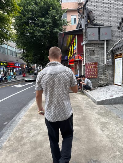A man walks away from the camera down a sidewalk in Liwan District, China. He is wearing a short-sleeved patterned shirt and black pants. In his right hand, he holds a small stack of bills with a red string tied around it. To his left, a street runs alongside the sidewalk with parked motorcycles, a silver car, and storefronts. To his right, a brick building with electrical boxes and signage stands adjacent to the sidewalk. A second man is seated on a raised platform in front of the brick building, looking at his phone. Overhead, trees and other buildings are visible. The lighting suggests it is daytime.