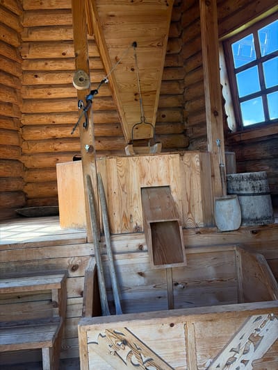 The image shows the interior of a wooden mill building located in the village of Kamaevo, Russia. The structure is constructed from logs, with visible wooden beams, planks, and stairs. Prominent features include a large wooden chute system, two long wooden poles leaning against a central support, a wooden shovel, and two large wooden barrels. A window with multiple panes allows light to enter, revealing a blue sky outside. The overall impression is of a rustic and functional mill, preserved with its original wooden elements.