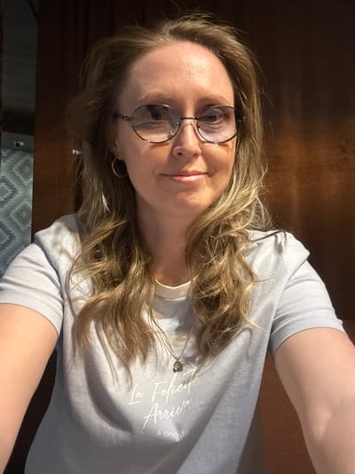 A woman with blonde, wavy hair and glasses is looking at the camera. She is wearing a light-colored t-shirt and a necklace with a small pendant. The background appears to be a wooden surface and part of a patterned blanket or throw. It is not possible to determine the location within Balezino, Russia, from the image.
