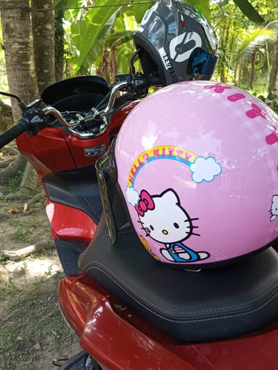 The scene depicts a red scooter parked outdoors, possibly in a residential area, given the lush green foliage and trees visible in the background. Two helmets rest on the scooter. A pink helmet decorated with Hello Kitty imagery sits on the scooter's seat. A black and grey helmet with the brand name "LEOPARD" is positioned on the handlebars. The scooter appears to be in a natural setting. Tanjongnie, Indonesia, cannot be confirmed.