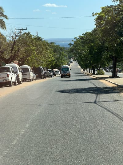 The image, taken from a slightly elevated perspective, shows a paved road curving gently to the right, lined on its left side by numerous green trees. The road surface is dark grey asphalt. Long, dark shadows of trees and utility poles stretch across the road from the left, indicating bright sunshine and possibly midday or early afternoon.

Along the left edge of the road, a line of various vehicles is parked or moving slowly. These include several white SUVs, one prominently displaying "Mahindra" on its rear, and a white commercial van with "NO. 70" and "Ozon" visible on its back. Further down the road, more cars, motorbikes, and a few people can be seen.

To the right of the road, the ground slopes down, revealing a vast, calm body of water—likely the Indian Ocean, given the provided location context of Pemba, Mozambique—stretching to the horizon under a clear, light blue sky. A few distant, light-colored clouds are visible. Utility poles with overhead wires run along the left side of the road, partially obscured by the tree canopy. The vegetation appears lush and tropical. The overall impression is a bustling street scene in a coastal town under bright, sunny conditions.