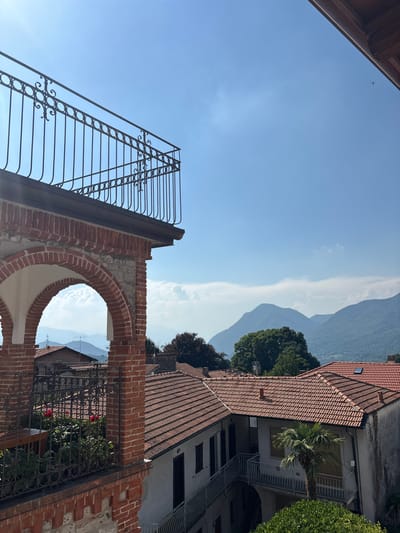 This media file is a vertical, medium-shot photograph taken outdoors during the day. The scene is a view through an arched brick opening, looking towards distant mountains.

The foreground features a portion of a brick structure with a large, semicircular archway. Through the arch, a black metal gate with an intricate scrolling pattern is visible, partially obscuring a lush green garden with several red roses. To the left of the arch, a black metal railing with decorative elements ascends diagonally against a clear blue sky. The sky is bright, suggesting a sunny day.

In the mid-ground, beyond the garden and gate, is a building with a red-tiled roof. Further in the background, a series of layered mountains rise against the sky. The atmosphere appears clear, with some distant haze or light clouds that create a soft contrast with the mountain slopes. The overall impression is one of a peaceful, scenic, and somewhat historic setting. There is no visible text in the image. The scene evokes a sense of tranquility and natural beauty, possibly in a rural or semi-rural location in Orino, Italy, given the architectural style and the presence of mountains and Mediterranean vegetation. The time of day is likely midday or early afternoon due to the bright sunlight and clear sky.