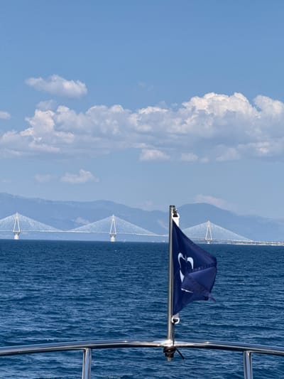 The image captures a scenic view of the Rio-Antirrio Bridge in Patras, Greece, from the perspective of a boat. The vast expanse of deep blue ocean dominates the right side of the frame, with gentle ripples suggesting a calm day. In the distance, the impressive cable-stayed bridge spans across the water, its white towers and cables stark against the hazy blue sky and distant mountains. The sky on the left is a bright, clear blue, punctuated by large, fluffy white clouds that cast soft shadows.
In the foreground, a dark blue flag with a white logo is affixed to a metal pole. The logo appears to be a stylized "3" or a similar abstract design. A metal railing runs along the right edge of the image, hinting at the boat's deck. A black rope is tied to the railing. The overall impression is one of a beautiful, sunny day, likely late morning or early afternoon, given the brightness of the sky. The scene evokes a sense of peace and the enjoyment of a maritime journey with a prominent landmark in the background. There is no visible text other than the logo on the flag.