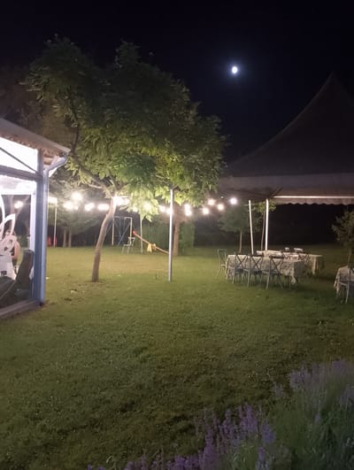 This media file depicts an outdoor dining area at night in Asparuhovo, Bulgaria. The scene is illuminated by string lights draped across trees and a large tented structure. Several tables are set with tablecloths and chairs, suggesting an event or gathering is being prepared or has recently concluded. In the background, a small playground with a swing set and a seesaw is visible. The moon is prominent in the dark sky. The overall atmosphere is serene and inviting, hinting at a festive or celebratory occasion.