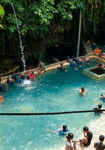 This image depicts a natural swimming pool with crystal-clear turquoise water surrounded by lush green vegetation and rocky cliffs. A small waterfall cascades into the pool from the upper left corner, creating a refreshing atmosphere. Several people are seen enjoying the water. In the foreground, a young woman in a white shirt and a young man in a black t-shirt are seated by the water's edge. In the middle ground, men and women are swimming and interacting in the pool. Some are gathered near the edge, while others are further out in the water. The pool has a stone border, and a submerged square structure is visible in the right section of the pool. In the background, more people are present, some sitting on steps leading down to the water, and others in the water. The overall scene suggests a popular recreational spot, likely a natural spring or waterfall, in Penungkiren, Indonesia, where visitors can relax and swim in a serene, natural environment.