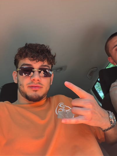 The image depicts the interior of a vehicle, presumed to be a car, in Sotirya, Bulgaria, during daylight hours. A male subject, appearing to be a young adult, is prominently featured in the foreground, facing the camera. He has dark, curly hair and a short beard and mustache. He is wearing a bright orange short-sleeved crew-neck shirt that features a partial white graphic design on the upper left chest/shoulder area. He wears dark, rectangular sunglasses with gold-colored accents; the lenses reflect what appears to be orange footwear and a blurred figure. His left hand (from the viewer's perspective) is raised, forming a gesture with the thumb and pinky finger extended and the middle three fingers curled. A metallic wristwatch with a dark strap is visible on his left wrist. A second male individual is partially visible in the background on the right, showing only the right side of his head with short, dark hair, his right ear, and a portion of his shoulder, clad in a dark garment over a lighter one. The car's light-colored, textured headliner is visible, along with a grey interior grab handle. A car window on the far right reveals green foliage outside. Strong, warm directional light, consistent with natural sunlight, illuminates the primary subject from the right.