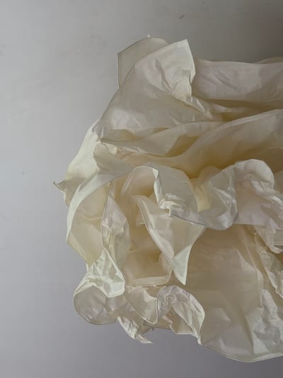 This is a close-up, overhead shot of a white, crumpled paper-like material. The material has been folded and arranged to create a voluminous, layered effect with many folds, creases, and soft curves. The texture appears to be thin and somewhat translucent, allowing light to pass through in some areas and creating shadows in others. The background is a plain, off-white surface, possibly a ceiling, with some subtle textural variations and a few small marks. The lighting is soft and diffused, highlighting the delicate nature of the material. The image is devoid of any people or discernible objects from a city environment. This image has no identifiable connection to Angelovo, Russia, as it depicts an abstract form rather than a specific scene or location.