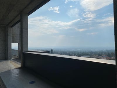 The image shows a view from a building overlooking the city of Kempton Park, South Africa, at coordinates -26.135 latitude, 28.201 longitude. The viewer is standing on a concrete balcony inside a building with a window to the left. The window is open to a concrete walkway. The view from the balcony is of the city below with a hazy sky above. The horizon is a line of trees and buildings, with the distant city skyline in the background. The sky is light blue with some white clouds. There are no people or objects in the scene other than the concrete structure of the building and the balcony.