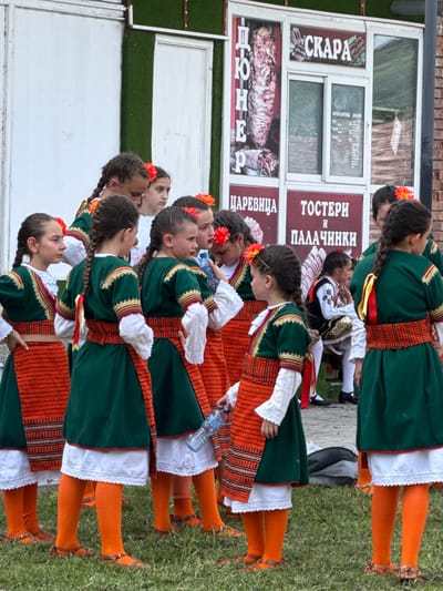 In Pordim, Bulgaria, a group of young girls dressed in traditional Bulgarian folk costumes stand in an outdoor setting. The costumes consist of green dresses with red and gold trim, paired with orange skirts, white blouses, and orange leggings. Many of the girls have their hair in braids adorned with red flowers. They are gathered in front of a building with signs in Bulgarian. One sign displays images of meat and the word "CKAPA", suggesting a food establishment. Another sign reads "ЦАРЕВИЦА TOCTEPH И ПАЛАЧИНКИ". The ground is covered in green grass. The overall atmosphere appears to be a cultural event or performance.