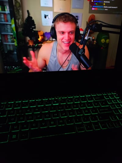 The image shows a person wearing headphones, looking at a computer screen and gesturing with their hands. The person is wearing a grey tank top and a gold chain. In the background, there are shelves filled with items, a small Christmas tree, and several framed pictures on the wall. There is also a green character figure on the right side of the screen. The lower half of the image shows a laptop keyboard with green backlighting. There is no indication of the city of Versailles, France, in this image.
