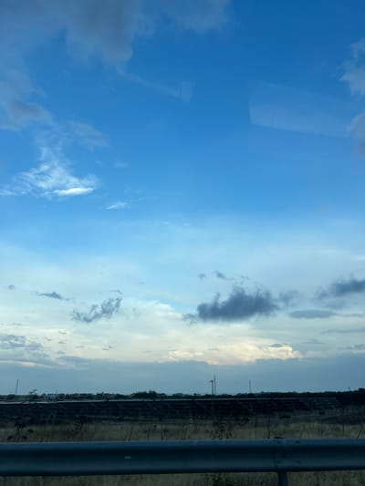 The image captures a wide shot of a sky filled with various cloud formations, ranging from wispy white clouds to darker, more ominous gray clouds. The sky transitions from a deep blue at the top to lighter shades of blue and white towards the horizon. Below the sky, a flat landscape is visible, characterized by dry, tall grass in the foreground. A dark, horizontal barrier, possibly a guardrail, runs across the lower portion of the frame. In the mid-ground, there is a large, dark structure that appears to be a solar panel farm, stretching across the scene. Beyond the solar farm, a line of buildings and some distant structures, including what looks like a power line tower, are visible. The overall impression is that of an overcast or partly cloudy day, viewed from a roadside setting. The location is identified as Manfredonia, Italy, suggesting the landscape and structures are characteristic of the region.