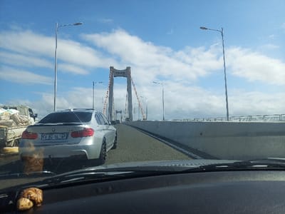 The image captures a bright, sunny daytime scene from the perspective of a vehicle moving across a large suspension bridge in Maputo, Mozambique, identified as the Maputo-Katembe Bridge. The dashboard and a small, turtle-shaped ornament are visible in the immediate foreground, looking through the windshield.

Ahead, a silver BMW sedan with the license plate "CV 80 VL GP" is driving in the right lane. To its left, partially visible, is a white truck loaded with several large sacks, suggesting it's carrying bulk materials like cement or sand. The road itself appears to be a multi-lane highway, bordered by concrete barriers and a metal guardrail on the right. Tall, grey streetlights with modern fixtures are spaced along the bridge.

In the midground and background, the prominent architectural features of the suspension bridge dominate. Its massive, reddish-orange and white pylons rise into the clear blue sky, supported by numerous suspension cables extending towards the road deck. Faint Chinese characters are visible on one of the pylons, likely identifying the construction company, "中国路桥" (China Road and Bridge Corporation), which built the bridge. The sky is clear with scattered white cumulus clouds, indicating good weather conditions. The scene depicts typical daytime traffic on this significant piece of infrastructure.