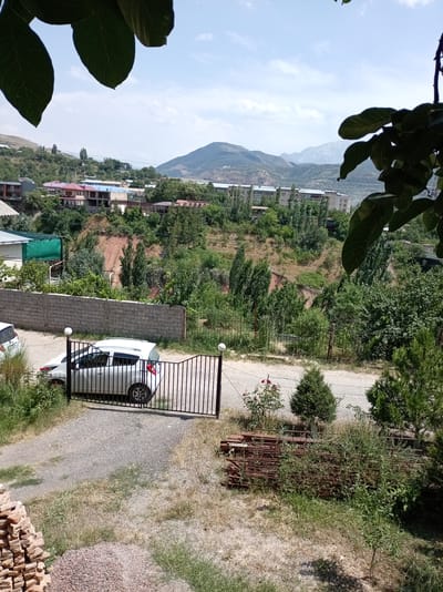 The image depicts a scene in the city of Charvak, Uzbekistan, during the daytime under a clear sky with scattered clouds. In the foreground, a gravel driveway leads to a black metal gate. A white hatchback car is parked just inside the gate. To the left of the driveway, a pile of bricks is visible. To the right of the gate, there is a stack of rusty metal rods overgrown with vegetation, and a small green tree. A low, grey brick wall runs horizontally across the middle ground, separating the driveway area from a grassy slope behind it.
Beyond the wall, a hillside is covered with trees and scattered buildings. The architecture of the buildings suggests a mix of residential and possibly some institutional structures, with various roof colors including red, blue, and grey. The hillside itself shows exposed earth in some areas, indicating natural erosion or construction activity. In the background, rolling hills and distant mountains form a scenic backdrop. The overall impression is of a semi-urban or suburban area nestled within a mountainous region. The foliage is lush and green, suggesting a warm season. The presence of leaves from a tree in the top left and right corners of the image frames the view, indicating the photo was taken from an elevated position, possibly from within another building or a balcony.