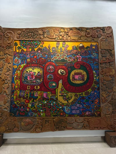 This is a close-up, eye-level shot of a vibrant, folk-art-style painting framed by a heavily carved wooden border. The painting itself is a chaotic yet engaging scene filled with numerous small figures, animals, and symbolic imagery. The dominant colors are bright yellow, red, blue, and green, creating a lively and energetic atmosphere.

The central focus of the painting appears to be a large, swirling red form, reminiscent of a stylized figure or an abstract representation of movement. Within this red form, and scattered throughout the painting, are circular vignettes depicting scenes of people in various domestic or social settings. One prominent circular scene shows a family gathered around a table. Other smaller circular windows reveal individuals and groups. Animals like fish, giraffes, octopuses, and turtles are interspersed throughout the composition, often rendered in a whimsical, child-like style. Trees laden with fruit and flowers add to the overall abundance and liveliness of the scene. The background is a mix of dark blue and yellow, with stylized representations of buildings and landscapes.

The wooden frame is intricately carved with a variety of motifs, including animals, human figures, and abstract patterns. These carvings add a three-dimensional texture and a sense of deep tradition to the artwork. The overall impression is one of joyful celebration and a rich tapestry of life, rendered with a unique and expressive artistic style. There is no clear indication of time of day, weather, or specific location within the painting itself, other than the context provided that it is from Rawa Burung, Indonesia. The emotions evoked are primarily positive, suggesting a sense of community, family, and the beauty of nature and life. No legible text is visible within the painting or its frame.