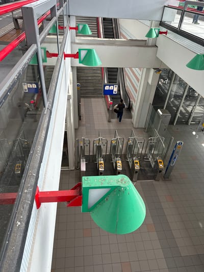 The photograph depicts the interior of a transit station in Lelystad, Netherlands. The focus is on the entry turnstiles and signage at the station's entrance. The turnstiles are arranged in rows with yellow card readers and a silver frame with glass barriers. Overhead signage displays the number "2" in blue lettering.
In the foreground, there is a row of green and red ticket reader posts situated at regular intervals. The reflections in the glass suggest the presence of an overhead structure. Tiles cover the walls, and a sign indicates directions to various platforms or locations within the station. Two individuals are walking towards an escalator in the distance. The environment has a modern architectural style, likely recently renovated or maintained.