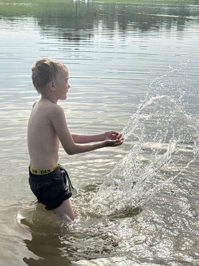 A fair-skinned, blonde-haired boy, wearing dark swim trunks with a yellow band imprinted with "D&N D&N," stands in a body of water, possibly a lake. He appears to be near the shore, based on the change in water color at the edge of the frame. The boy's arms are raised, with water spraying in an arc around them. Trees are visible in the background. The city of Novosinkovo, Russia, cannot be confirmed as the location from the image alone.