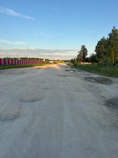 This is a photograph of a dirt road in Izyayu, Russia. The road is in disrepair with numerous potholes and puddles, indicating it may have recently rained or is poorly maintained. A red and grey striped metal fence runs along the left side of the road. Beyond the fence, there are trees and some structures. Streetlights are visible along the road. The sky is blue with some wispy clouds, and the sun appears to be low in the sky, casting long shadows. The overall scene suggests a rural or semi-rural environment.