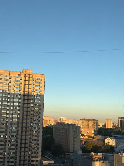 The image depicts a cityscape under a clear blue sky, likely during the golden hour as evidenced by the warm sunlight illuminating the buildings. In the foreground, a tall, multi-story brick building dominates the left side of the frame. Its facade is a patchwork of windows, with some reflecting the sunlight and others in shadow. The building's design appears to be functional, possibly residential or office space.

Further into the background, a collection of other buildings of varying heights and architectural styles are visible. These include mid-rise apartment buildings with orange-toned exteriors, as well as some lighter-colored structures. Trees dot the urban landscape, suggesting a degree of green space interspersed with the concrete and brick. A thin, dark horizontal line, possibly a wire or cable, stretches across the upper portion of the sky. The overall impression is one of a densely populated urban environment at the end of the day.