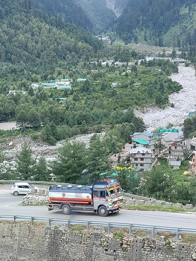 This image depicts a mountainous landscape in Palchan, India, with a winding road carrying traffic. In the foreground, a tanker truck, likely carrying fuel, drives on the right side of the road. The truck is silver with a white tank that features orange and blue accents and has logos and writing. A white car is visible in the background, also on the road. The road is bordered by a stone wall on the left and a metal guardrail on the right. The background is characterized by lush green, densely forested mountainsides that rise steeply. Scattered throughout the hillside are buildings with light-colored walls and predominantly green or gray roofs, suggesting a village or small town nestled within the mountains. A dry, rocky riverbed or debris flow is visible in the upper right portion of the image, indicating the terrain. The overall scene suggests a remote, possibly rural, mountainous area with ongoing transportation activity.