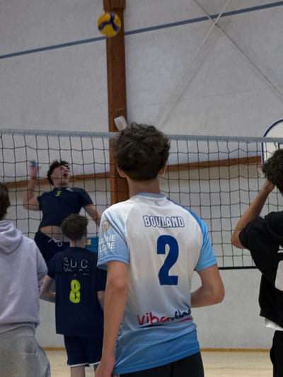 An indoor volleyball game is captured in an athletic facility located in Sennecey-le-Grand, France. The primary subject in the immediate foreground, viewed from the back, is a male individual wearing a white and light blue short-sleeved jersey with "BOULAND" printed above the large blue numeral "2". His hair is dark and curly.
Mid-ground, beyond a black volleyball net, another male individual, dressed in a dark blue t-shirt, is airborne, mid-jump, with his mouth open and right arm raised, positioned to interact with the volleyball. A yellow and blue volleyball is suspended in mid-air, slightly above and to the left of the airborne player, over the net. To the left of the airborne player, also behind the net and facing away, is another male wearing a dark blue t-shirt with "S.U.C." and a yellow numeral "8" visible on the back. Partially visible to the far left is a male in a light purple or grey hooded garment, also facing away. A partial view of another individual with dark curly hair is visible to the right of the foreground player.
The environment consists of a light-colored court floor and light-colored walls or ceiling, with a visible brown wooden structural beam on the right side of the frame and white horizontal lines on the wall. The volleyball net spans the width of the playing area.