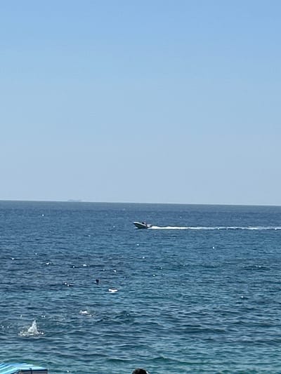 The image depicts a wide expanse of blue ocean under a clear blue sky. In the middle ground, a white speedboat is visible, moving from left to right, leaving a white wake behind it. Further in the distance, on the horizon, a faint, dark shape suggests the presence of land or a larger vessel. Closer to the viewer, in the foreground, the water is a deeper blue with numerous small white reflections or splashes. A hint of a striped beach chair is visible in the bottom left corner, and a dark head is just visible in the bottom center, suggesting people are present at the edge of the water. The overall scene conveys a calm, sunny day at a coastal location.