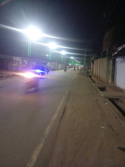 The image depicts a street scene at night in Tundun Wada West, Nigeria. The road is illuminated by bright streetlights, some with green accents. On the left side of the road, there are buildings with shops and what appear to be fences. A motorcycle with its headlights on is moving down the road, creating a bright, blurred effect due to motion. Several people are visible walking along the roadside and on the street. The right side of the road features a sidewalk or dirt path, a drainage ditch with concrete coverings, and more buildings. The overall atmosphere is that of a busy street at night, with activity and illumination.