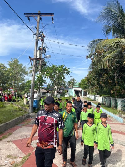 In Beuringen, Indonesia, under a bright blue sky with scattered clouds, a group of people walks down a paved pathway lined with vegetation. The group appears to be mostly male, with many younger individuals wearing green long-sleeved shirts and black skullcaps, suggesting they may be students or attending a religious event. An adult male in the foreground wears a red and black patterned shirt with "1945 INDEPENDENCE DAY" printed on it. He is walking towards the camera, looking slightly to the side. Behind him, other adults and children are visible, some dressed in similar green attire, others in different clothing. A prominent utility pole with electrical wires and a transformer stands to the left of the pathway. In the background, there are buildings, trees with lush foliage, and a white fence. The scene depicts a bright, daytime outdoor gathering or procession in a semi-urban or village environment.