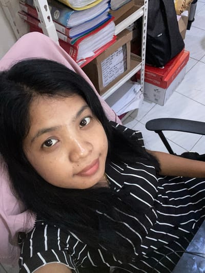 The image is a close-up selfie of a person with dark hair and tan skin, wearing a black and white striped shirt. The person is looking at the camera with a slight smile. The background is a cluttered workspace with shelves containing files, boxes, and other items. The environment appears to be indoors with tiled walls. The location is potentially Negara, Indonesia.