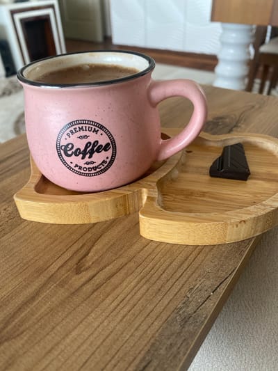 An overhead shot of a pink speckled coffee mug filled with coffee sits on a wooden tray. The mug has "Premium Coffee Products" printed on it in black ink. Next to the mug, on the same tray, is a piece of dark chocolate. The tray is shaped like a heart. The tray is placed on a wooden table. The background is blurred but shows what appears to be a living room with framed art on the wall. The image was taken in Talas, Turkey.