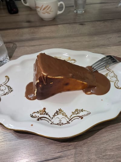 A slice of chocolate-covered cheesecake sits on a decorative white plate with gold trim and ornate brown floral designs. A silver fork is partially inserted into the cheesecake. The plate is placed on a wooden table. In the background, two white mugs with a similar floral design are visible, as well as a glass of water and a remote control. The scene appears to be indoors. This image does not contain any information related to the city of Yalangach, Uzbekistan.