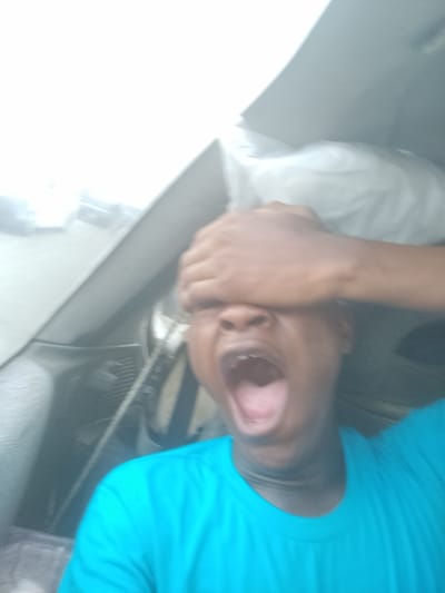 A man with dark skin is inside a vehicle, covering his eyes with his hand and yelling. He is wearing a blue shirt. The interior of the car is visible, including the dashboard and a white object possibly covered in plastic wrap on the back seat. Outside the car window, there are blurry images suggesting an urban environment, likely Ilishan, Nigeria.