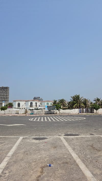 This full-shot image captures a sunny outdoor scene, likely in Bac Cam Ranh, Vietnam. The perspective is slightly elevated, looking across a street towards a row of buildings and the sky.

In the foreground, a wide asphalt road or parking area spans the lower and right portions of the frame. It features clear white painted markings, including solid lines for lanes or parking spaces, and a distinctive semicircular white striped median or turn lane marking on the left. The pavement appears somewhat worn and dirty, with a discarded plastic water bottle with a blue label visible on the far right.

The midground, stretching along the left and center, showcases a series of light-colored buildings, predominantly white, with red-tiled roofs, suggestive of Mediterranean or resort architecture. These structures are nestled among abundant green foliage, including numerous palm trees that line the street. Further down the street on the left, an unfinished concrete high-rise building is visible.

The background is dominated by a vast, clear, light blue sky, indicating bright, fair weather. There is no discernible human activity or specific event taking place, giving the scene a quiet and still atmosphere. The time of day appears to be mid-day or early afternoon due to the bright, shadow-less light. The text on the water bottle label is illegible due to distance and blur.