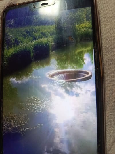 A photograph on a smartphone screen displays a scenic view of a body of water with lush green vegetation and trees along its banks. The water surface reflects the sky and surrounding foliage, creating a serene and natural ambiance. In the center of the water, a circular structure, possibly a manhole or a well cover, is visible. The image is taken from a slightly tilted angle, suggesting it might be a photo taken by someone holding the phone. The location within the city of Svoboda, Russia, is not discernable from the visual information presented in the image.
