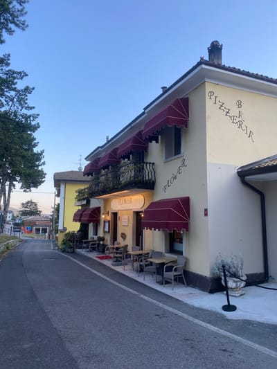 The image shows a light yellow building with a sign that says "FLOWER" and "PIZZERIA" on it, located in the city of Caprino Veronese, Italy. The building has red awnings over windows and doors, and outdoor seating with tables and chairs. There is a paved road in front of the building, and a tree on the left side of the image. The sky is clear and blue. The scene appears to be set during daytime, with no people visible in the image.