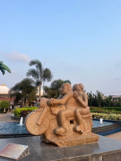 This image shows a stone sculpture of two human figures on a bicycle, installed in a water fountain. The sculpture has a rough texture and appears to be weathered. The background features palm trees, various other plants, and a building in the distance. The sky above is light blue. The base of the sculpture is black granite. A plaque is attached to the base.

The plaque contains the following text:

ariu Gopal
id his work
snifuenced by Anna Hazare.
Corruption in the countr some
Supported by the
ton ycles where hundreds
Remotorcycle procession and
soudarity for the cause.
PRAKALP GOA