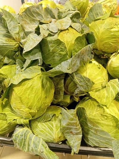 The image shows a close-up view of a pile of green cabbages in a market stall. The cabbages are arranged in a haphazard manner, with some leaves overlapping others. The cabbages are mostly whole, with some being cut in half, and their surfaces display a range of green hues, with some leaves appearing slightly brown. The cabbages are likely located in a market stall within the city of Pridelands, Kenya, based on the provided coordinates.