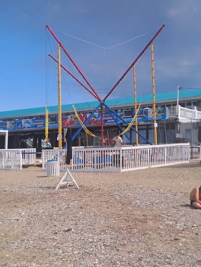 A wide shot captures a bungee trampoline attraction situated on a pebble beach in Dzhubga, Russia. The attraction consists of a tall, red, V-shaped frame supported by two yellow poles. Multiple elastic cords are attached to the frame, leading down to harnesses that are currently empty. The scene is set against a backdrop of a light blue building with a teal roof. In the foreground, a white picket fence encloses the trampoline area, and a few people are visible in the background. One person, a male wearing a light-colored shirt, is standing near the attraction, while another, a young boy, is sitting on the beach to the right. The beach itself is covered in small, grey pebbles and shells. The sky is a mix of blue and dark clouds.