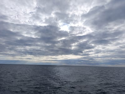 The image depicts a wide expanse of the ocean under a cloudy sky. The water is dark and choppy, with small waves visible. A streak of sunlight reflects off the water in the middle distance. The sky is filled with a thick layer of grey and white clouds, with a few patches of brighter sky showing through in the upper center of the image. The horizon line is clearly visible, separating the sea from the sky. The scene appears to be a natural, open environment, likely at sea, with no man-made structures or people visible. The location is identified as Unknown, Sweden, suggesting a geographical context of a coastal or maritime area within the country.