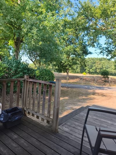 The image depicts a wooden deck with a railing on the left side, overlooking a sun-drenched landscape. A black trash bag is visible on the deck, placed in a dark-colored bin. In the foreground, a portion of an outdoor chair is present. Beyond the deck, a gravel path leads into a dry, grassy field dotted with trees. In the distance, a dense forest borders the field, under a clear blue sky. The scene appears to be an outdoor seating area, likely in a park or campsite, given the rustic wooden structures and the natural surroundings. The lighting suggests it is daytime, with the sun casting shadows.