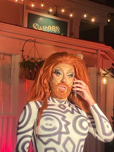 A drag performer with voluminous red hair, heavy stage makeup, and a ginger beard is on the phone, holding a green cell phone to their right ear. They are wearing a white, long-sleeved bodysuit adorned with a bold, abstract pattern of dark blue concentric circles and angular shapes. The performer's expression is one of surprise or concern.
In the background, a building with white siding is visible. Above the building, a sign with the word "SAILORS" in stylized yellow lettering against a dark green background is illuminated. A string of small, glowing light bulbs runs across the top of the image, casting a warm light. To the left of the performer, a hanging basket filled with green plants and pink flowers is suspended from a metal hook. The overall lighting suggests it is nighttime, with warm tones and some red-tinted ambient light. The setting appears to be an outdoor area of a establishment in Provincetown, United States of America, possibly a bar or performance venue.