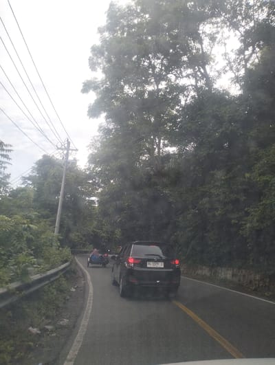 The image depicts a view from a vehicle traveling on a narrow, winding asphalt road lined with guardrails and dense foliage. The road has a yellow center line and a white edge line. In the distance, a motorcycle with a sidecar carrying two people is visible. A black car with a license plate that reads "BL 1179 J" is in the foreground, also on the road. Power lines run overhead, supported by a concrete utility pole. The environment appears to be a tropical or subtropical area, with lush green trees and vegetation on either side of the road. The sky is overcast. The location is identified as Seungkomulat, Indonesia.
