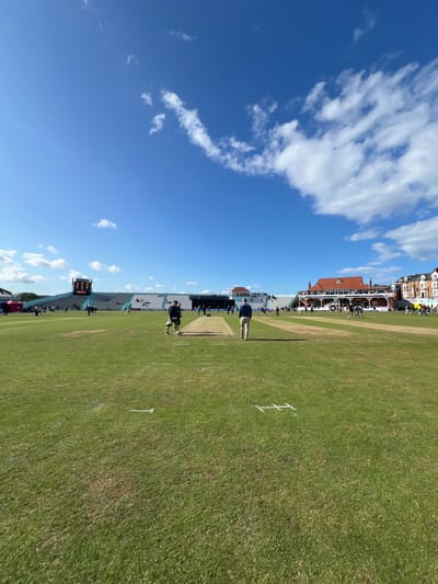 This image depicts a wide-angle view of a cricket ground, identified as North Marine Road Ground in Scarborough, United Kingdom, under a bright, clear blue sky with scattered white clouds.
In the foreground, a manicured green grass outfield is visible, marked with faint white lines, possibly indicating field boundaries or pitch measurements. Two male figures are centrally positioned in the midground, walking away from the viewer towards the cricket pitch. The figure on the left wears a dark shirt and light trousers; the figure on the right wears a blue jacket and light trousers. Further down the pitch, additional individuals, possibly players or ground staff, are present, some appearing to be engaged in pre-match preparations or light activity.
The background features the stadium infrastructure. To the left, a large electronic scoreboard displays numerical information. Adjacent to the scoreboard and extending across the left-center, are extensive, tiered spectator stands, predominantly light blue in color, with sections populated by spectators. To the right, a prominent, traditional-style pavilion building with a distinctive red-tiled roof, white facade, and multiple balconies is visible, with people present on its levels. Beyond the pavilion to the far right, a row of multi-story buildings, consistent with traditional British terraced architecture, lines the perimeter of the ground. The scene indicates a daytime event, likely a cricket match or practice session, with clear weather conditions.