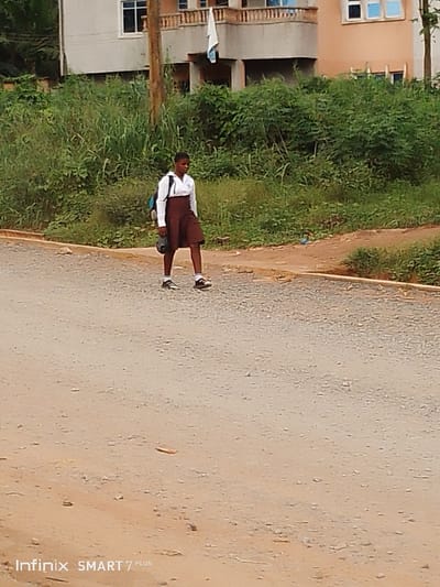A young woman, likely a student, walks along a dusty road. She wears a school uniform consisting of a white top and a brown skirt, with a backpack slung over her shoulder. Lush green vegetation lines the side of the road and a building is in the background. The lighting indicates it's daytime, and the setting appears to be a rural or suburban area. The road itself is unpaved and uneven.