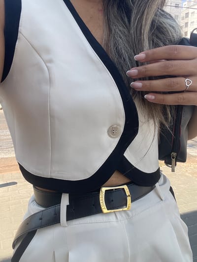 The image displays a partial view of a person, likely female, from the upper chest to the mid-thigh, captured outdoors in an urban setting within Anápolis, Brazil. The person is wearing a sleeveless, cropped vest that is primarily cream/off-white with contrasting black trim along the neckline, armholes, and hem. A single translucent button with a subtle swirled pattern is visible on the right side of the vest. Below the vest, a black belt with a textured surface secures a pair of cream/off-white pleated pants or a skirt. The belt features a rectangular gold buckle, vertically engraved with the text "J. DARROY" on both the left and right sides.

The person's right hand is visible on the right side of the frame, adorned with a silver-colored ring featuring an open heart design on the ring finger. The fingernails are manicured with a French tip style. Long, light brown hair with visible highlights cascades over the person's left shoulder and chest. A black bag or purse with a visible zipper pull is partially seen on the left side of the image, behind the person's arm.

The ground beneath the person consists of a paved surface, appearing to be a sidewalk constructed from reddish-brown interlocking concrete pavers. The background is softly blurred, indicating distant urban structures under bright daylight conditions.