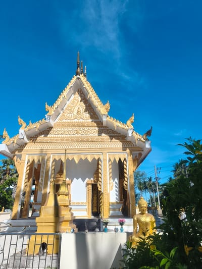 A golden Buddhist temple with ornate carvings stands against a bright blue sky in Tong Yang, Thailand. The temple's roof is adorned with decorative finials and intricate patterns, and its facade features white walls with golden accents. In front of the temple, a golden Buddha statue is seated amongst lush green foliage. The scene is illuminated by clear daylight, indicating a sunny day.