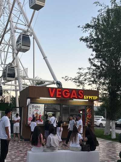 In Manas, Kyrgyzstan, a vibrant scene unfolds at what appears to be an amusement or recreational area. Dominating the view is a large, white Ferris wheel, its intricate structure and enclosed gondolas hinting at an exhilarating experience. Adjacent to the Ferris wheel is a food stall, its facade adorned with a prominent neon sign that reads "VEGAS" in large, glowing red letters, with the word "БУРГЕР" (Burger) in yellow lettering above it. The stall also features eye-catching advertisements for burgers and "Bubble Tea."
Several people are gathered around the food stall, some ordering, others waiting to receive their food, and some are seen walking by. The atmosphere suggests a casual, lively gathering. The time of day appears to be late afternoon or early evening, with the sky a soft blue and the lighting casting a warm glow.
The overall setting is an outdoor public space, likely a park or entertainment venue, with trees providing a natural backdrop and paved pathways indicating a well-maintained area. The people are dressed casually, reflecting a relaxed outdoor environment. The presence of a Ferris wheel and food vendors points to a place designed for leisure and enjoyment.