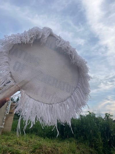 The image, taken in Aghalokpe, Nigeria, features a large, circular textile object, predominantly white with a dense, long white fringe around its entire perimeter. The object appears to be a ceremonial fan or a decorative hand fan, as a fabric-wrapped handle is visible at its base. A hand with dark skin is seen in the lower right corner, holding this handle, positioning the object upwards against the sky and natural surroundings.

The main surface of the white textile has a textured appearance and shows faint, light grey or blue patterning that is not discernible as specific text or symbols. The background reveals an outdoor setting on what appears to be a cloudy day, providing soft, diffused lighting. The upper and left portions of the background show a light blue sky with scattered white clouds. The right and lower parts of the background are filled with lush, green vegetation, consistent with tall grass or dense bushes. A small section of a concrete building structure is also visible near the bottom right, behind the foliage. The perspective of the photograph looks upwards, emphasizing the fringed object against the natural backdrop. No specific activity beyond holding the object is depicted, and no emotions are discernible.