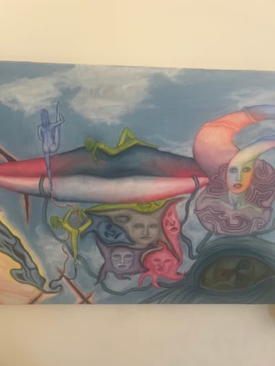 This is an oil painting on canvas depicting a surreal and abstract composition. The background is a light blue sky with wispy white clouds. The central subject is a large, elongated, amorphous shape with bands of red, white, and grey.
Several stylized human figures are incorporated into the scene. A blue figure is contorted in a yoga-like pose, suspended from the upper left. A green figure reclines on top of the central amorphous shape, also in a relaxed pose. Another green figure is in a similar contorted pose below and to the left of the central shape.
Scattered across the lower portion of the painting are several faces and mask-like forms. There are at least four distinct faces, rendered in shades of grey, purple, and pink, some with open mouths. One pink form resembles an octopus with a face.
To the right, a large, distorted, colorful head emerges. This head has a pale blue face with yellow and orange accents around the eyes and nose. It has a voluminous, wavy hairdo in shades of purple. The head appears to be emerging from a large, pinkish-orange, curved form that extends towards the top right of the canvas.
In the bottom right corner, a large, dark, eye-like shape is visible, filled with abstract forms.
The painting is presented in a horizontal format. There is no discernible location in the city of Hadera, Israel, mentioned or depicted within the artwork itself. The scene is fantastical and not representative of any real-world setting.