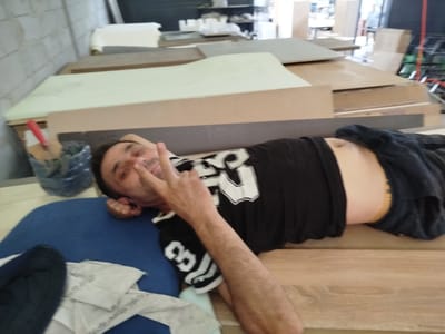 A man is lying on a blue surface, with his head resting on his hand. He is wearing a black t-shirt with white graphics and dark blue shorts. His facial expression is difficult to ascertain due to the angle and resolution of the image, but he appears to be looking at the camera. In the background, there are various stacks of wooden boards and some industrial equipment, suggesting a workshop or storage area. The overall environment appears to be indoors, with a somewhat cluttered and unfinished look. The image does not provide any specific landmarks or clues that would allow for pinpointing a location in Burgalik, Uzbekistan.